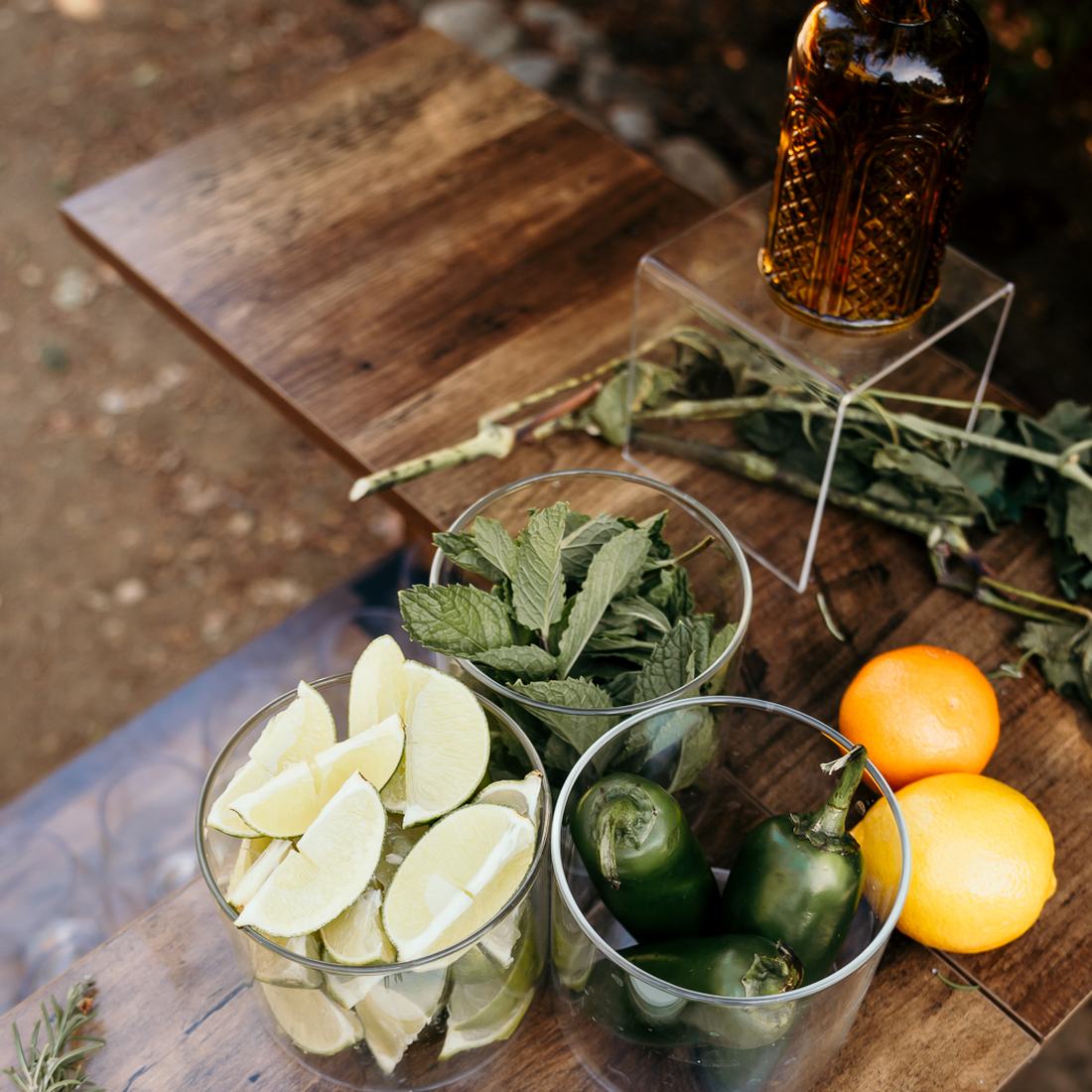 Glass vessels of lime, mint and jalapenos beside oranges and lemons on a wooden bar